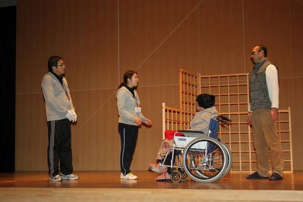 舞台の上で車いすに乗った女性の後ろにいる男性と話をしている介護職員2名の写真